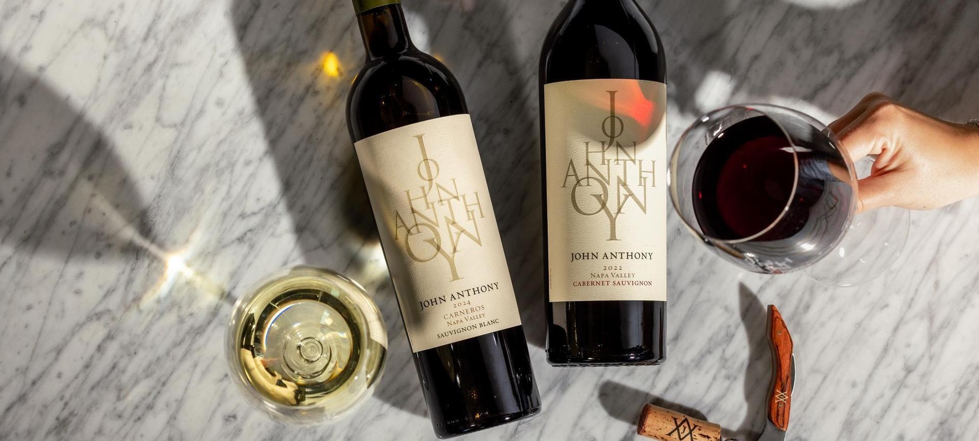An overhead shot of two John Anthony wine bottles—a Sauvignon Blanc and a Cabernet Sauvignon—resting on a marble surface with two glasses of wine.