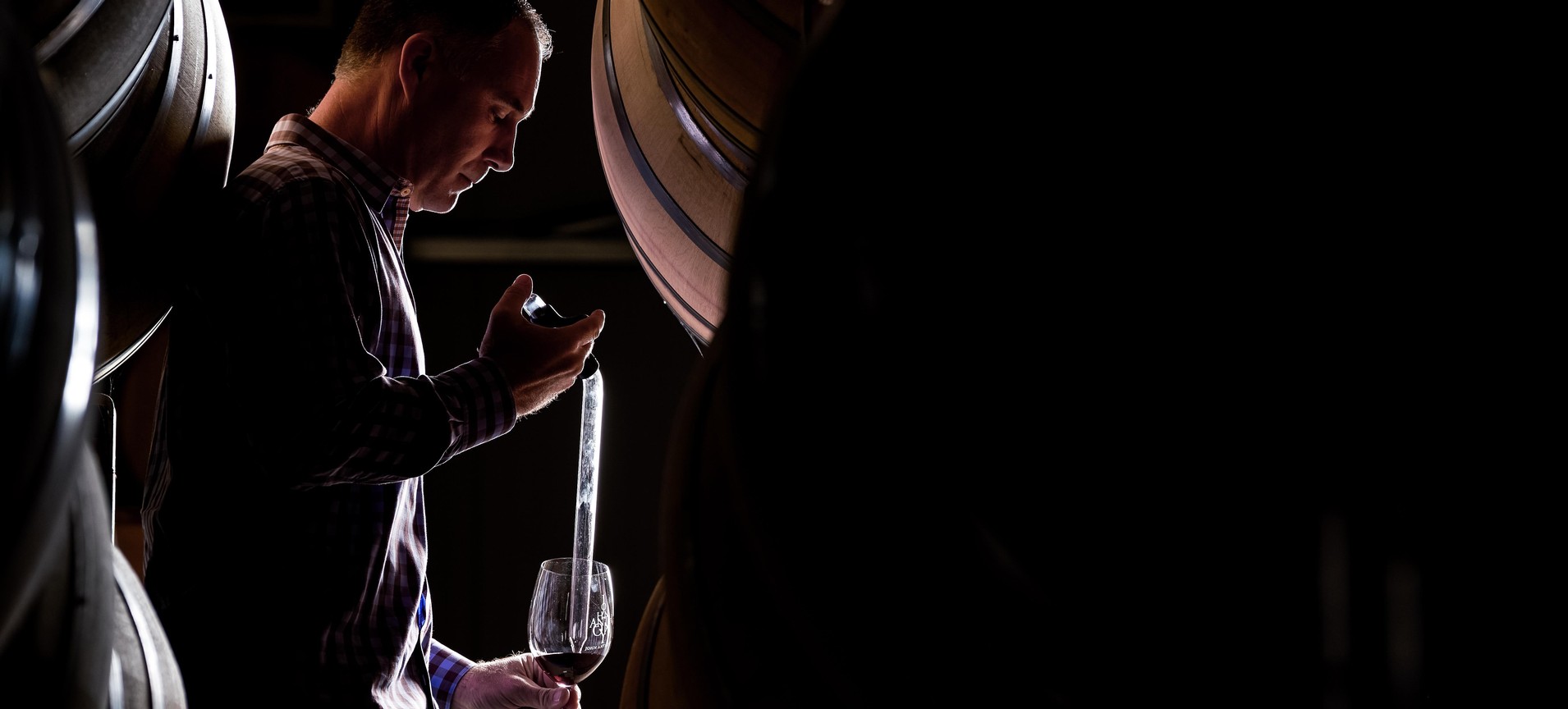 A person in a dark cellar uses a wine thief to draw wine from a wooden barrel into a glass, captured in dramatic low-light.